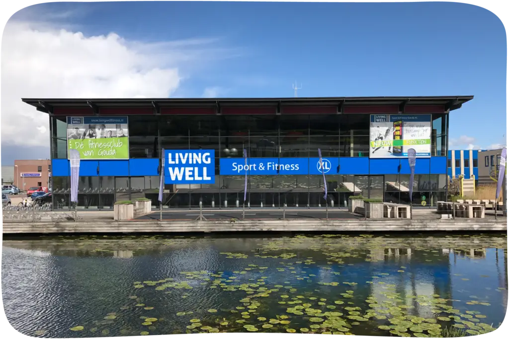 Recht vooraanzicht van het grote fitnesscentrum Living Well Sport & Fitness XL in Gouda met felle blauwe gevelbelettering en grote outdoor spanframes vanaf het water gezien.