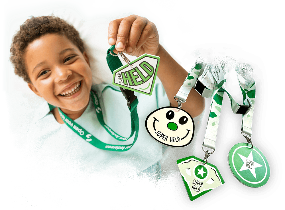 Een vrolijk glimlachend kind in een ziekenhuisbed dat een groene heldenmedaille met de tekst Super Held omhooghoudt, met daarnaast een overzicht van verschillende ontwerpen heldenmedailles aan keycords.
