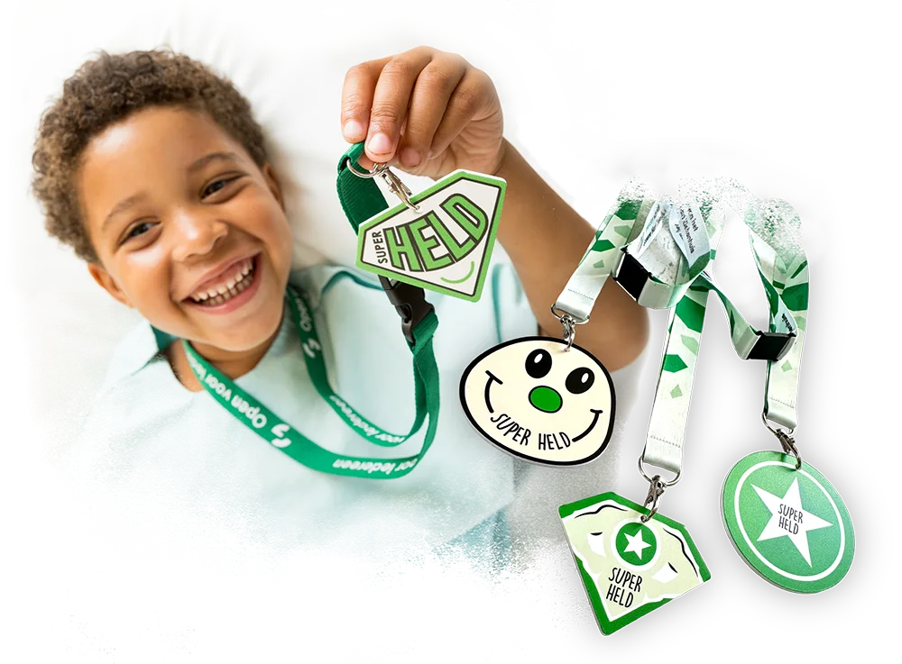 Een vrolijk glimlachend kind in een ziekenhuisbed dat een groene heldenmedaille met de tekst Super Held omhooghoudt, met daarnaast een overzicht van verschillende ontwerpen heldenmedailles aan keycords.