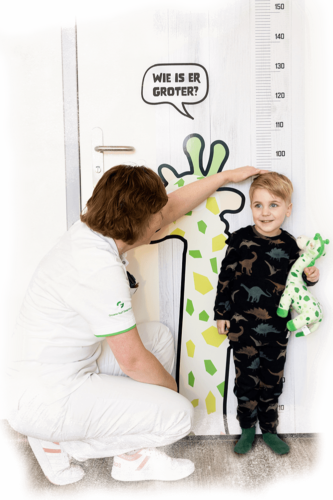 Een verpleegkundige die de lengte opmeet van een glimlachend jongetje bij een grote deurbestickering van een meetlat met Freek de Giraffe en de tekst Wie is er groter.