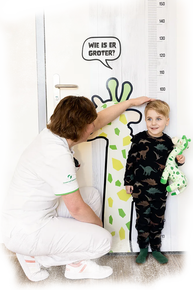 Een verpleegkundige die de lengte opmeet van een glimlachend jongetje bij een grote deurbestickering van een meetlat met Freek de Giraffe en de tekst Wie is er groter.